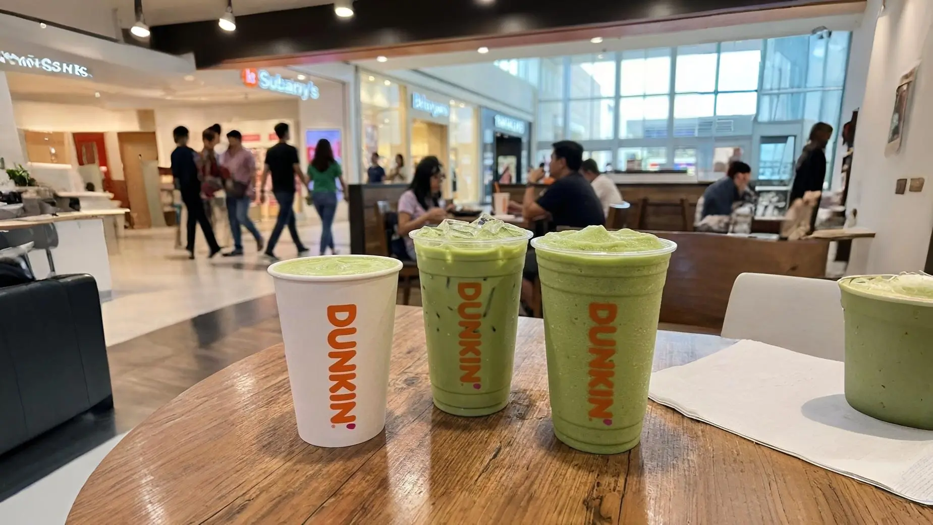 Dunkin matcha drinks including hot, iced and frozen matcha lattes displayed on a wooden table inside a busy shopping mall cafe
